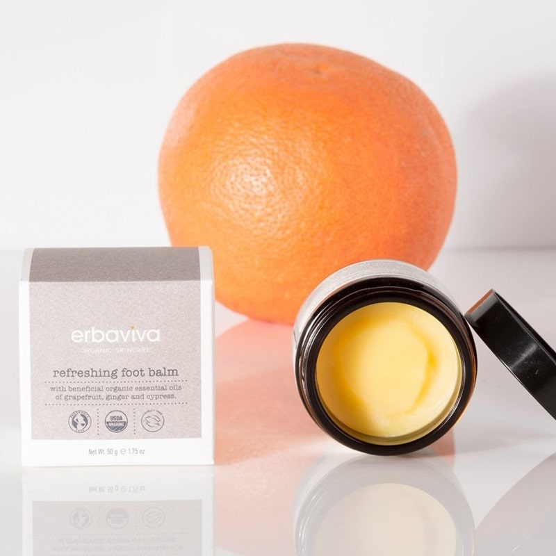 Lifestyle shot of Erbaviva Refreshing Organic Foot Balm shown with lid off (1.75 oz) with box and orange in the background