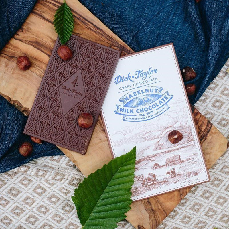 Dick Taylor Craft Chocolate Hazelnut Milk Chocolate - product shown next to packaging and hazelnuts on wood plank