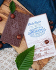 Dick Taylor Craft Chocolate Hazelnut Milk Chocolate - product shown next to packaging and hazelnuts on wood plank