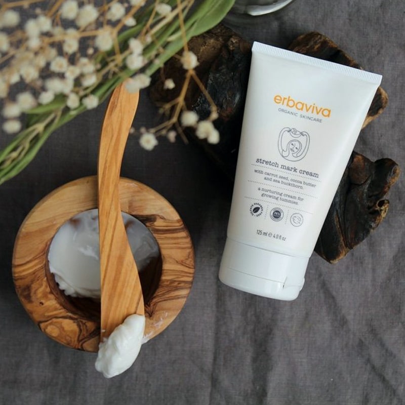 Lifestyle shot of Erbaviva Stretch Mark Cream (4 oz) with product shown in wood bowl and product on end of wood spoon and delicate white flowers in the background