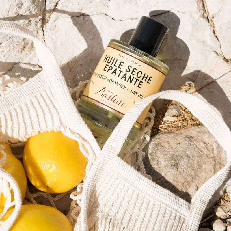 Lifestyle shot of Bastide Huile D&#39;Aix Orange Blossom Bath Oil (250 ml) shown top view with sack of lemons in the foreground