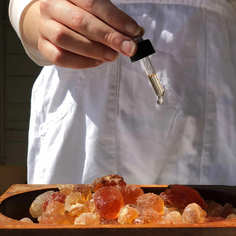 Lifestyle shot of model holding dropper of Bastide Ambre d&#39;Or Potpourri Refresher Oil over Ambre d&#39;Or Potpourri Crystals