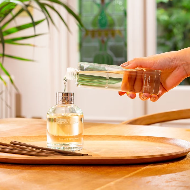 Lifestyle shot of Molton Brown Orange & Bergamot Aroma Reeds Diffuser Refill (150 ml) shown being poured into diffuser