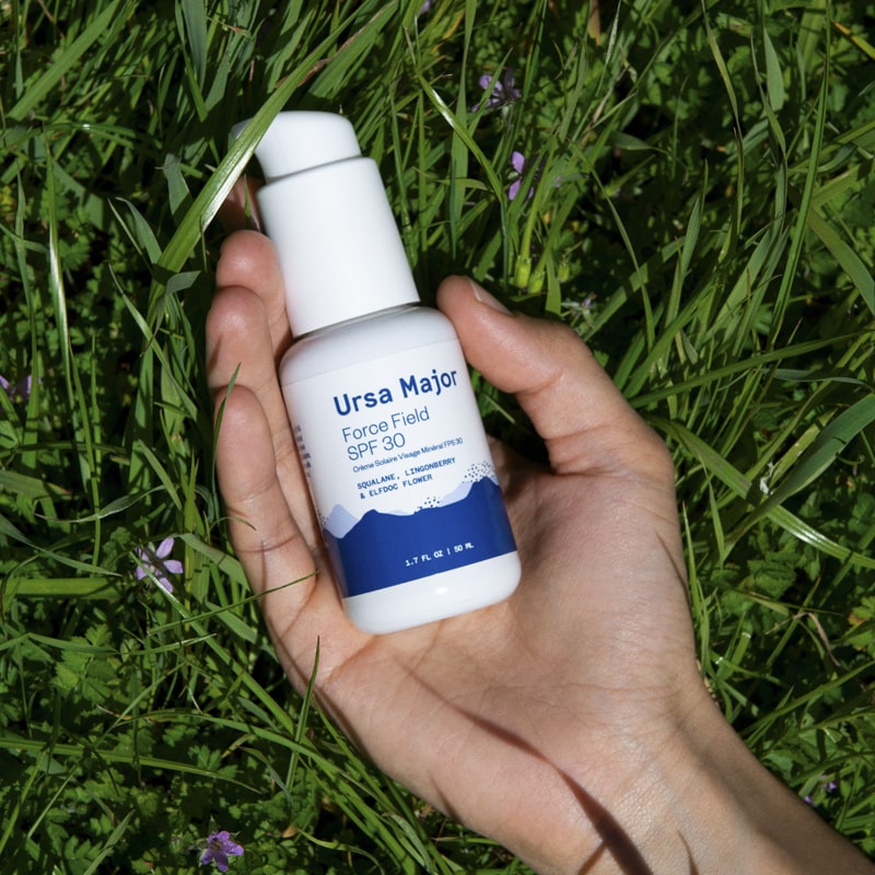 Lifestyle shot of Ursa Major Force Field SPF 30 shown in hand of model with green grass and purple flowers in the background