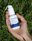 Lifestyle shot of Ursa Major Force Field SPF 30 shown in hand of model with green grass and purple flowers in the background