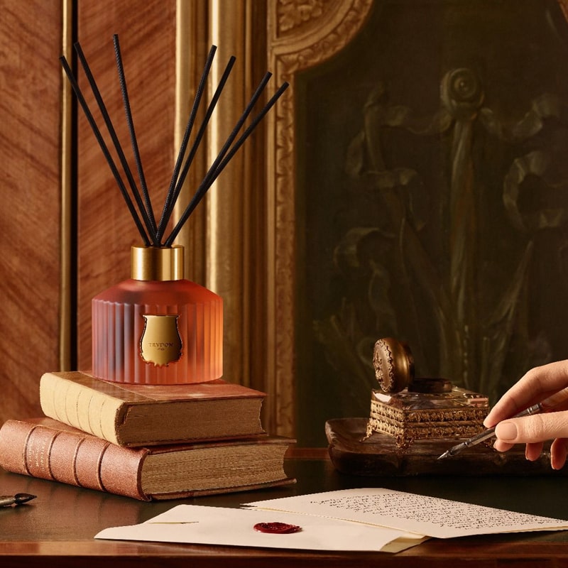 Lifestyle shot of Trudon Tuileries Home Fragrance Diffuser (350 ml) sitting on a stack of books on a desk, with a person writting a letter with a calligraphy pen in the foreground