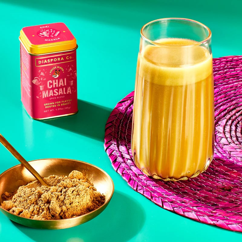 Lifestyle shot of Diaspora Co Chai Masala (1.59 oz) shown in tin with gold bowl showing Chai Masala powder and Chai Masala drink made in glass