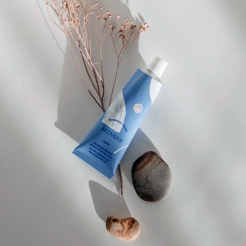 Lifestyle shot - top view of Minois Paris Pommade Fesses (Diaper Cream) (50 ml) tube with rocks and dried flowers in the background