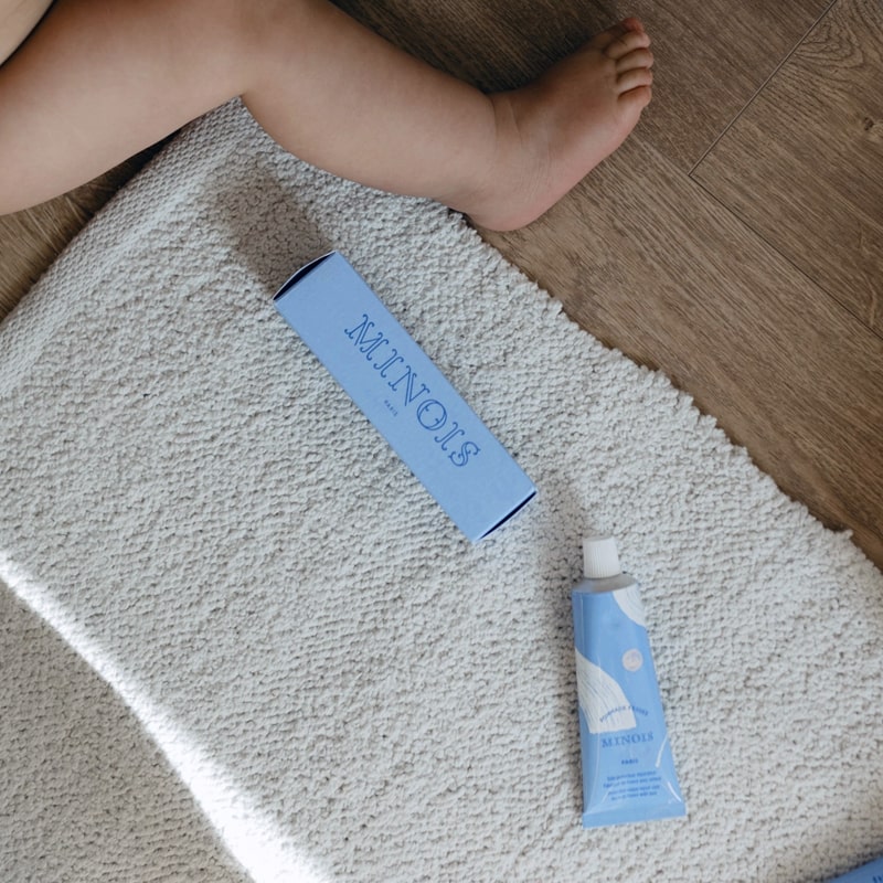 Lifestyle shot - close up of Minois Paris Pommade Fesses (Diaper Cream) (50 ml) tube and box on rug with leg of baby shown