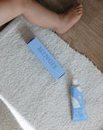 Lifestyle shot - close up of Minois Paris Pommade Fesses (Diaper Cream) (50 ml) tube and box on rug with leg of baby shown