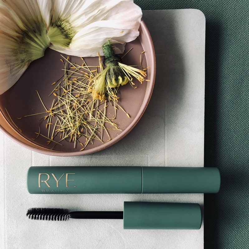 Lifestyle shot top view of Rye Beauty Brow Grooming Serum (7 ml) showing wand next to closed tube and flower pieces in the background