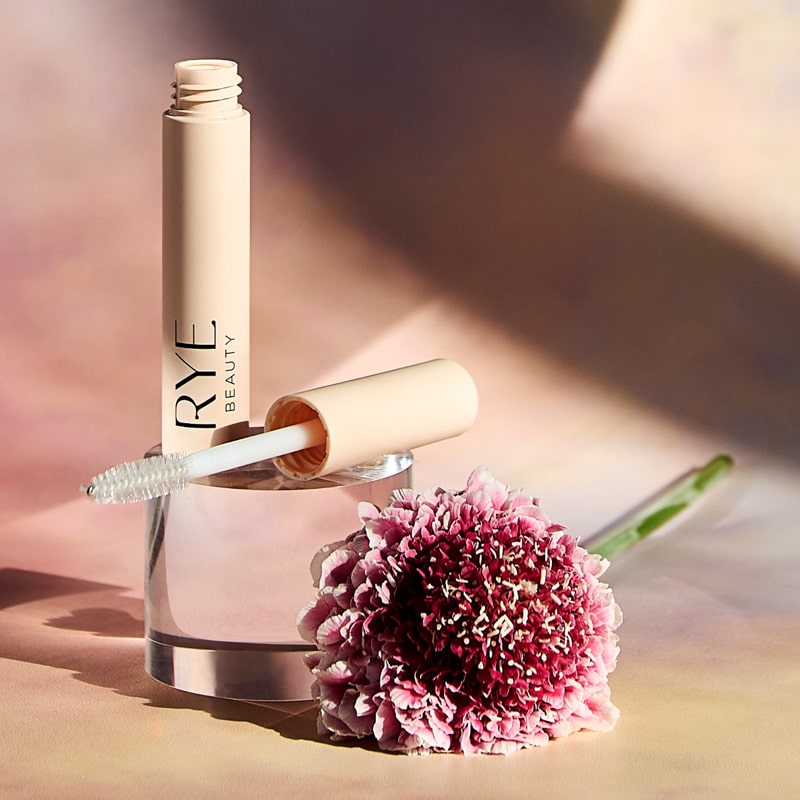 Lifestyle shot of Rye Beauty Brow Milk (6 ml) tube shown open with wand and pink flower in the foreground