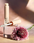 Lifestyle shot of Rye Beauty Brow Milk (6 ml) tube shown open with wand and pink flower in the foreground