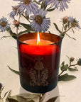 The Quiet Botanist In The Weeds Scented Candle - lit candle shown in front of flowers