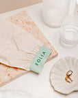 Folia Body Exfoliating Tan Removal Mitt - product shown on sink next to glasses, stone slab, and earrings