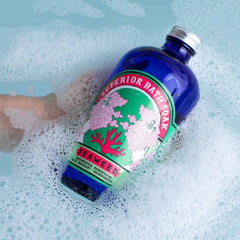 Lifestyle shot of Archivist Seaweed Bath Soak (480 ml) shown in hand of model in tub of water and suds