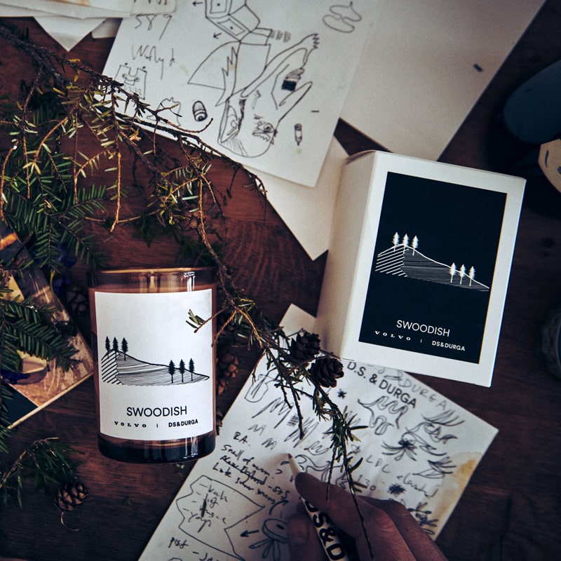 Lifestyle shot top view of D.S. &amp; Durga Swoodish Candle (7 oz) and box with pine needles and drawings in the background