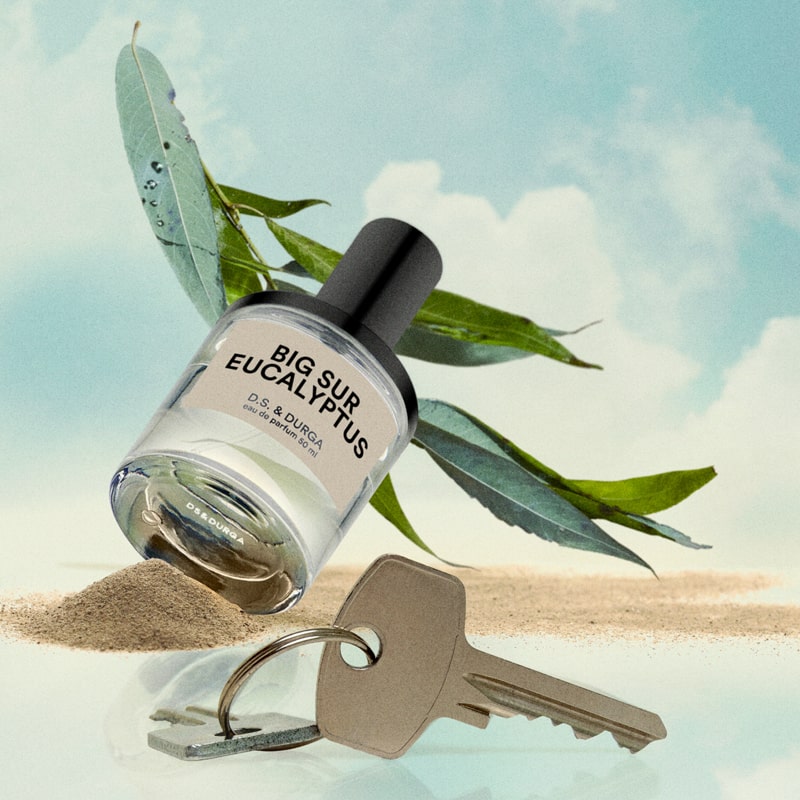 Lifestyle shot of Big Sur Eucalyptus Eau de Parfum (50 ml) with keys on ring in the foreground with sand and eucalyptus in the background with blue sky and clouds.