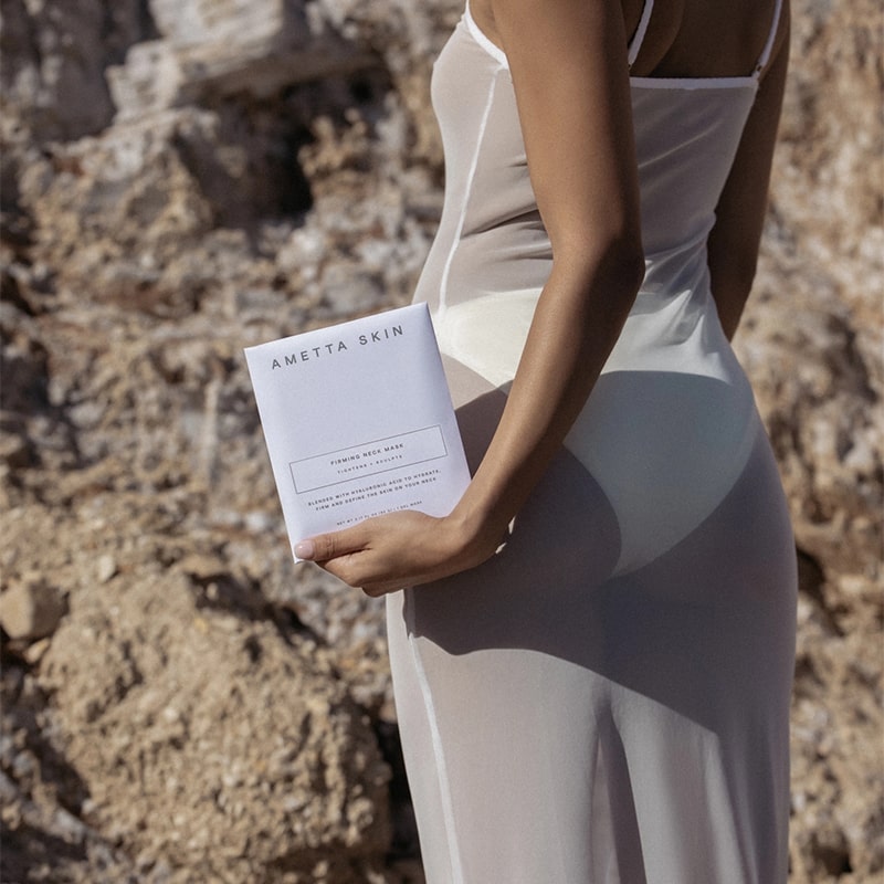 Ametta Skin Firming Neck Mask - model shown holding product packaging on beach