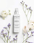 Lifestyle shot top view of Leonor Greyl Complexe Energisant - Leave-In Invigorating Scalp Treatment For Thinning Hair (150 ml) with purple flowers andn white roses placed around the bottle