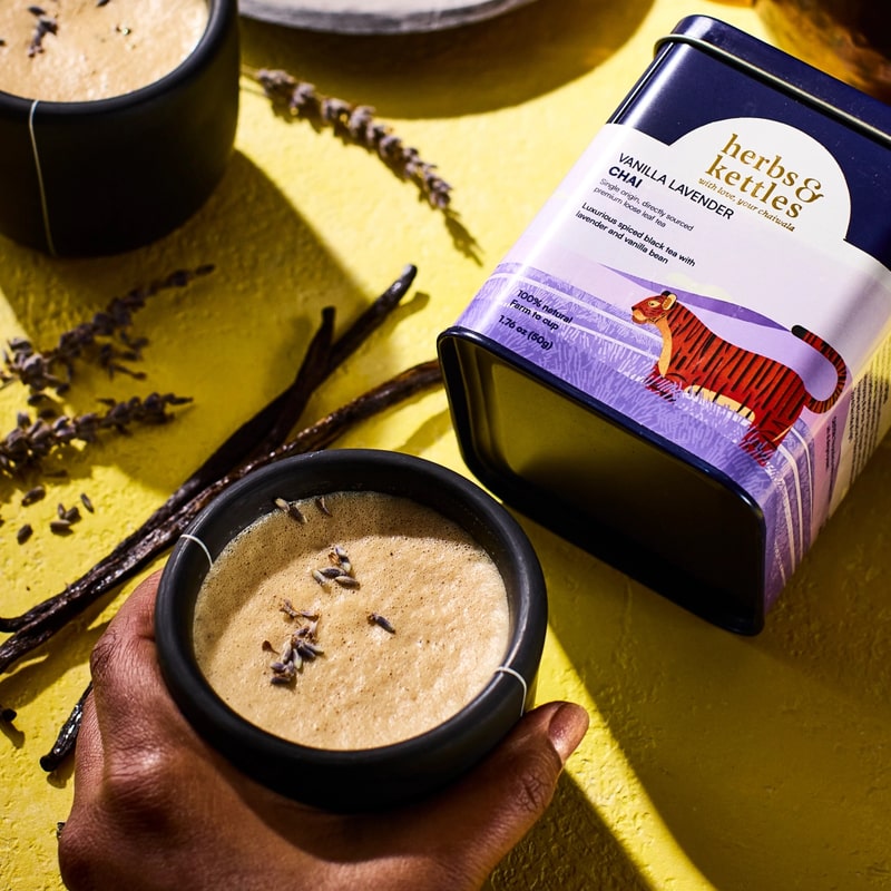 Lifestyle shot of Herbs & Kettles Vanilla Lavender Chai (1.76 oz) shown prepared in ceramic mug with vanilla bean pods and lavender in the background with tin