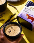 Lifestyle shot of Herbs & Kettles Vanilla Lavender Chai (1.76 oz) shown prepared in ceramic mug with vanilla bean pods and lavender in the background with tin