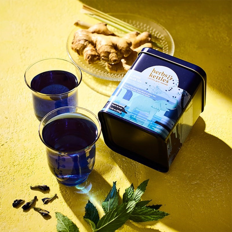 Herbs & Kettles Lavish Blue Tea - product packaging shown next to plate of ginger and herbs with two glasses of tea