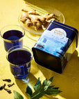 Herbs & Kettles Lavish Blue Tea - product packaging shown next to plate of ginger and herbs with two glasses of tea