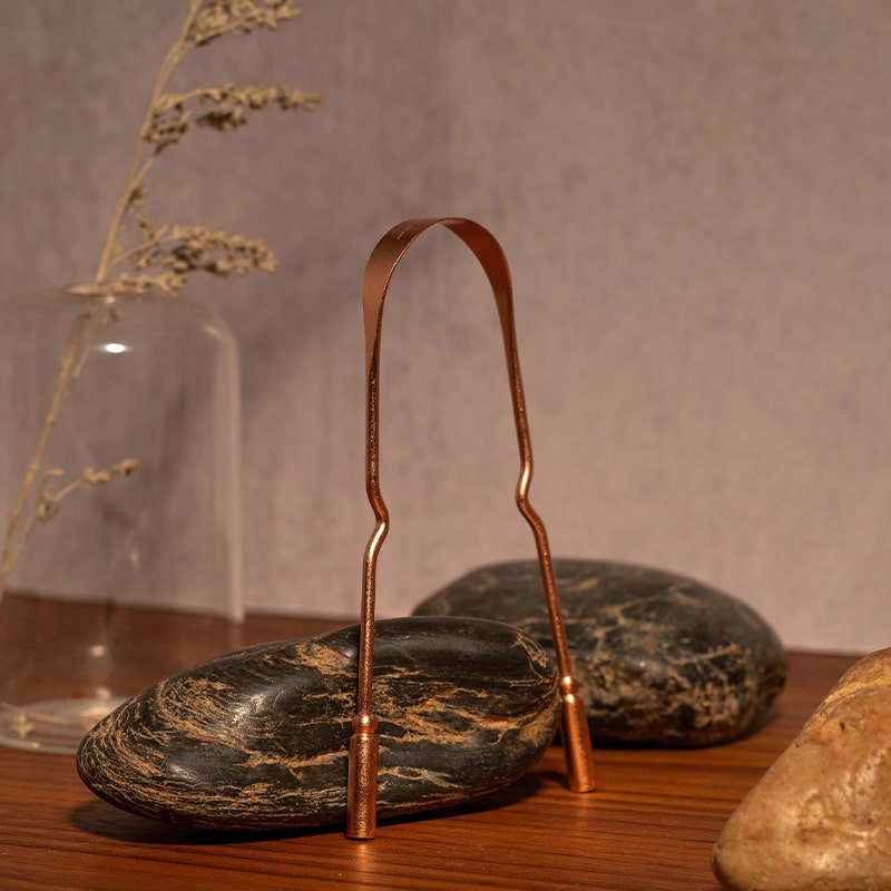 KOPPEN Pure Copper Tongue Scraper - product shown next to stones and glass vase with plant