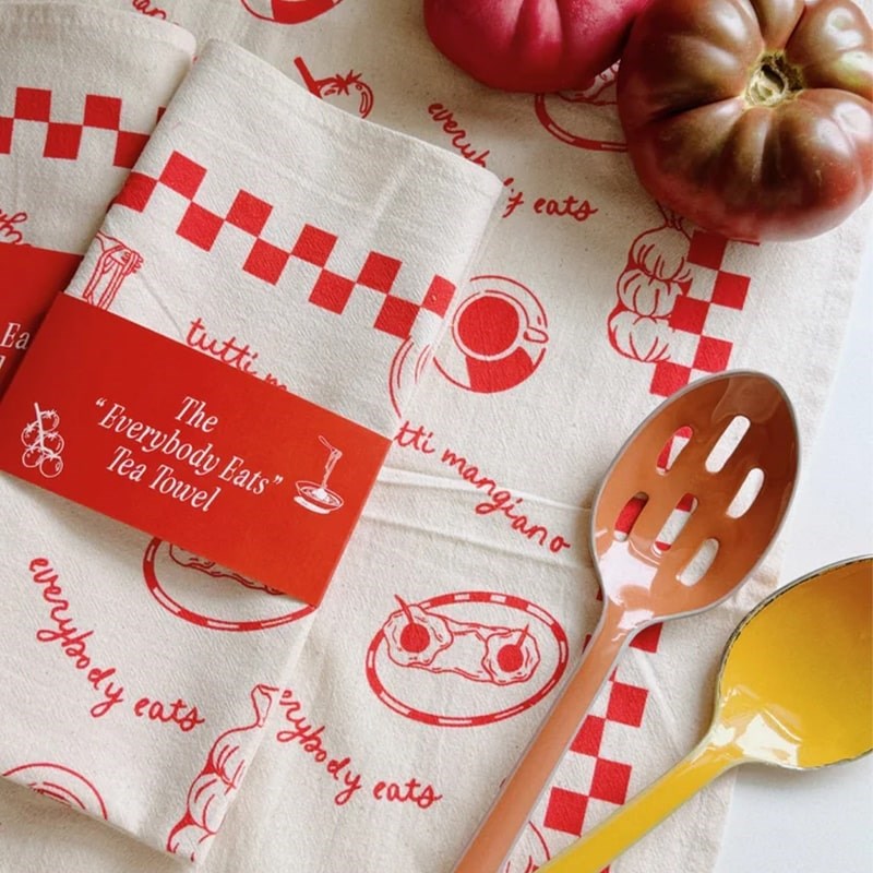 Mel Andrel Everybody Eats Tea Towel - folded product next to tomatoes and cooking spoon