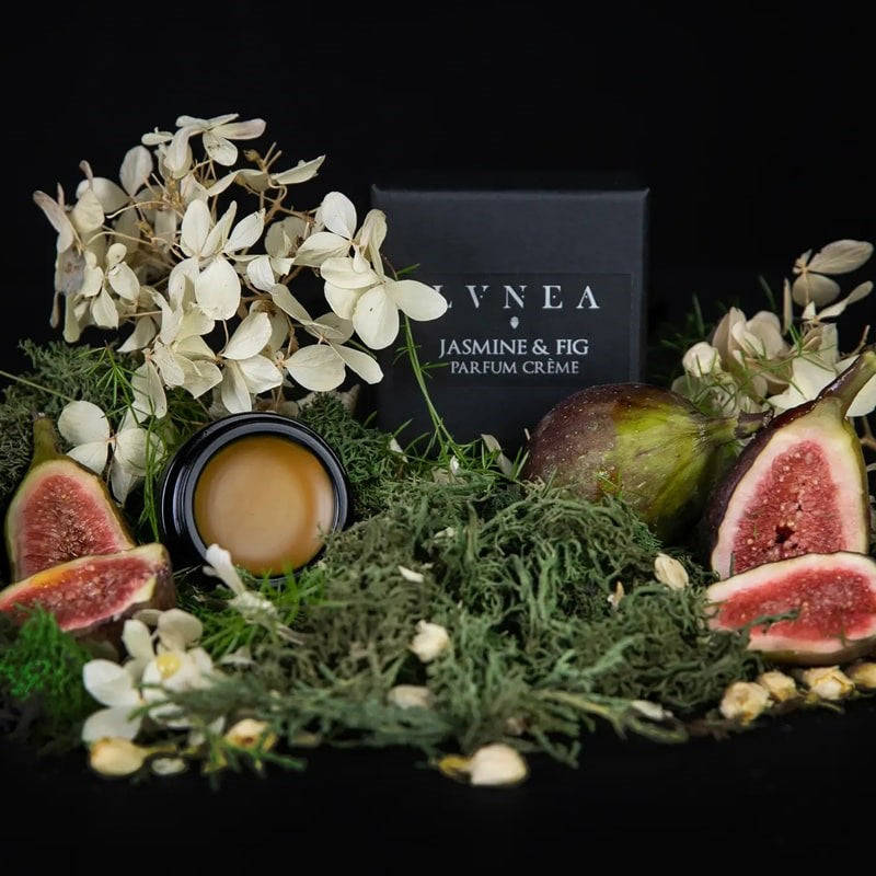 Lifestyle shot of Lvnea Perfume Jasmine & Fig Parfum Creme (10 g) box and opened jar with figs in the foreground and white delicate flowers