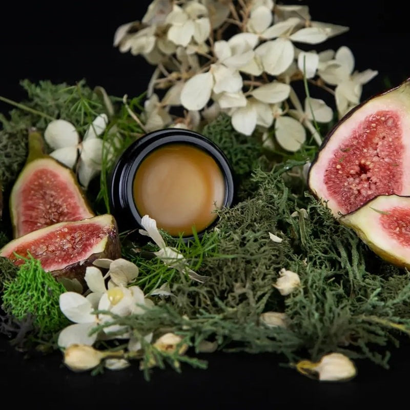 Lifestyle shot of Lvnea Perfume Jasmine & Fig Parfum Creme (10 g) opened jar so show color of product with figs in the foreground and white delicate flowers